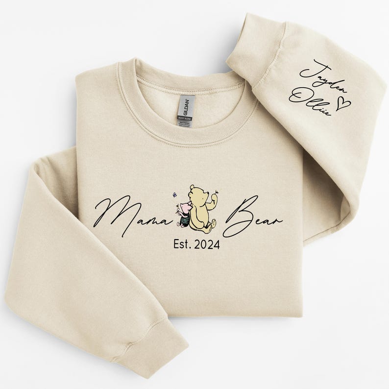 May include: A beige sweatshirt with the text "Mama Bear Est. 2024" and a cartoon image of Winnie the Pooh and Piglet. The right sleeve has the text "Taylor Ollie" with a heart.