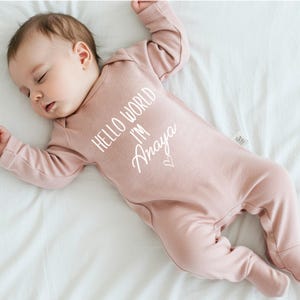 Pode incluir: Um macac&atilde;o rosa de manga comprida para beb&ecirc; com o texto "Hello World I'm Anaya" em branco.