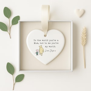 K&ouml;nnte beinhalten: Ein herzf&ouml;rmiger Keramikschmuck mit cremefarbenem Band. Das Ornament zeigt eine Illustration von Winnie Puuh und Ferkel sowie den Text "To the world you're a Mum, but to me you're my world. Love Josie x."