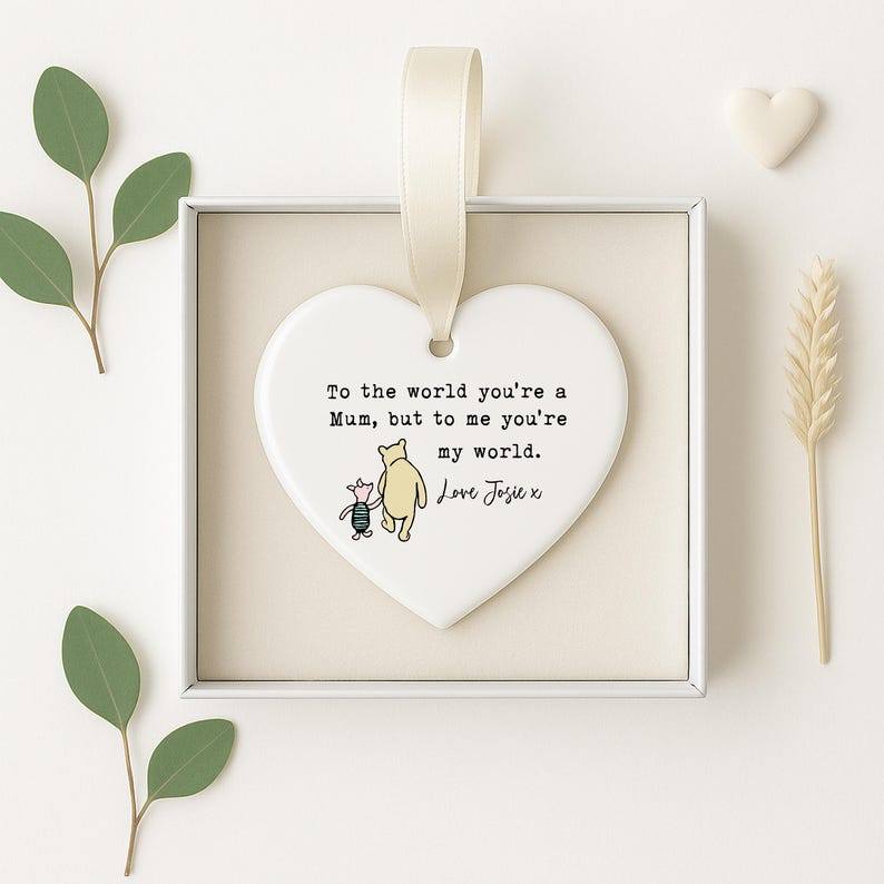 K&ouml;nnte beinhalten: Ein herzf&ouml;rmiger Keramikschmuck mit cremefarbenem Band. Das Ornament zeigt eine Illustration von Winnie Puuh und Ferkel sowie den Text "To the world you're a Mum, but to me you're my world. Love Josie x."