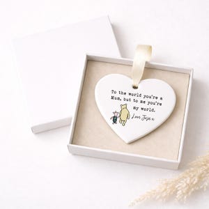 K&ouml;nnte beinhalten: Wei&szlig;es herzf&ouml;rmiges Ornament mit cremefarbenem Band, pr&auml;sentiert in einer wei&szlig;en Box. Das Ornament zeigt eine Illustration von Winnie Puuh und Ferkel mit dem Text: "To the world you're a Mum, but to me you're my world. Love Josie x."