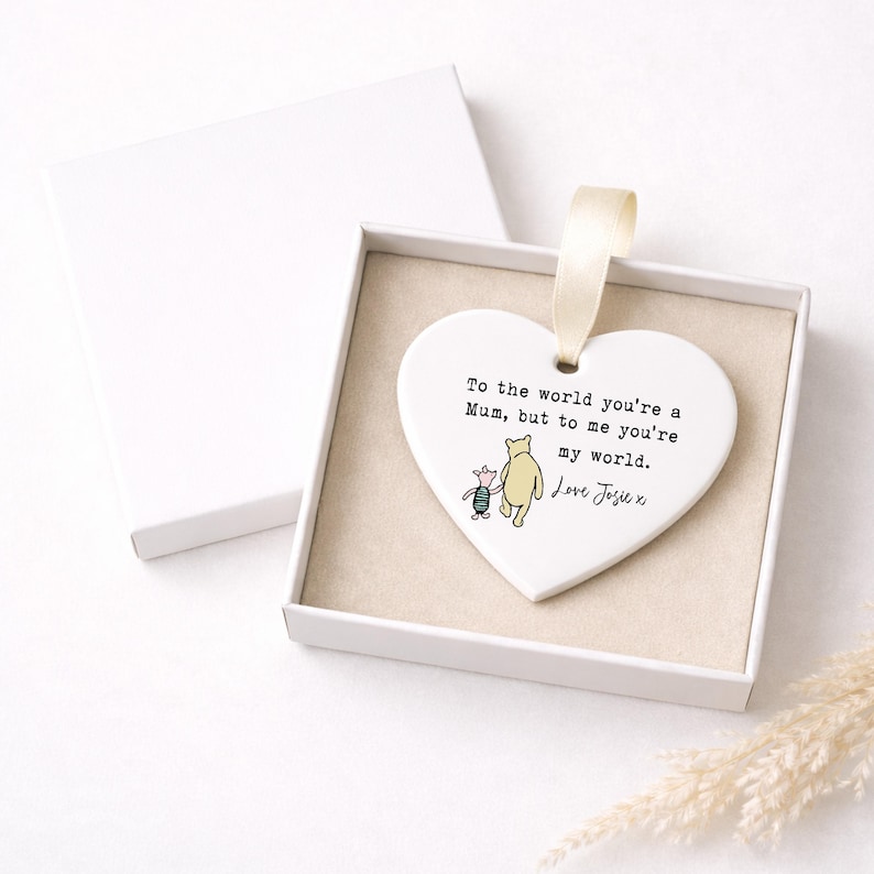 K&ouml;nnte beinhalten: Wei&szlig;es herzf&ouml;rmiges Ornament mit cremefarbenem Band, pr&auml;sentiert in einer wei&szlig;en Box. Das Ornament zeigt eine Illustration von Winnie Puuh und Ferkel mit dem Text: "To the world you're a Mum, but to me you're my world. Love Josie x."