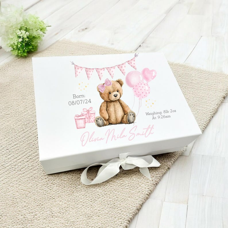 May include: White keepsake box with a pink and brown teddy bear illustration, pink balloons, and gift boxes. The box has a pink banner and the text "Born: 08/07/24, Weighing: 8lb 2oz At 9:26am" and the name "Olivia Mila Smith". A white ribbon is tied around the box.