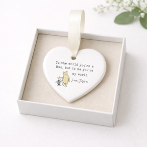 K&ouml;nnte beinhalten: Wei&szlig;es herzf&ouml;rmiges Ornament mit cremefarbenem Band, in einer wei&szlig;en Schachtel. Das Ornament zeigt eine Winnie-Puuh-Illustration und den Text: "To the world you're a Mum, but to me you're my world. Love Josie x."