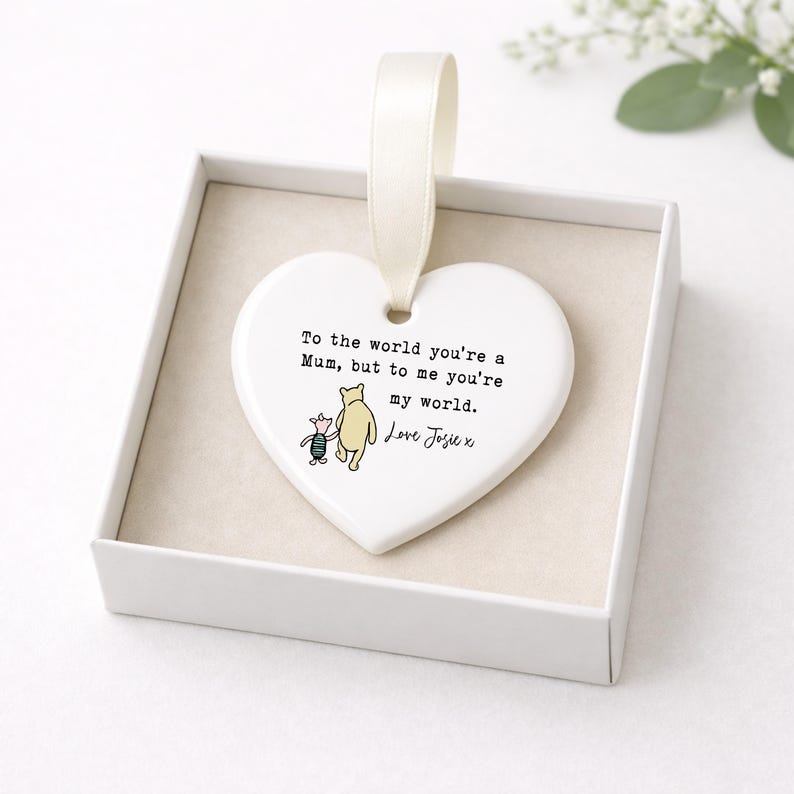 K&ouml;nnte beinhalten: Wei&szlig;es herzf&ouml;rmiges Ornament mit cremefarbenem Band, in einer wei&szlig;en Schachtel. Das Ornament zeigt eine Winnie-Puuh-Illustration und den Text: "To the world you're a Mum, but to me you're my world. Love Josie x."