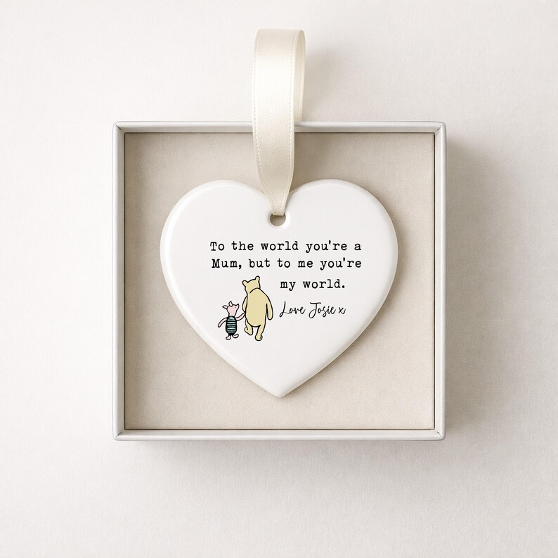 K&ouml;nnte beinhalten: Wei&szlig;es herzf&ouml;rmiges Keramikornament mit cremefarbenem Band. Es zeigt eine Winnie-the-Pooh-Illustration und den Text: &bdquo;To the world you're a Mum, but to me you're my world. Love Josie x.&ldquo;