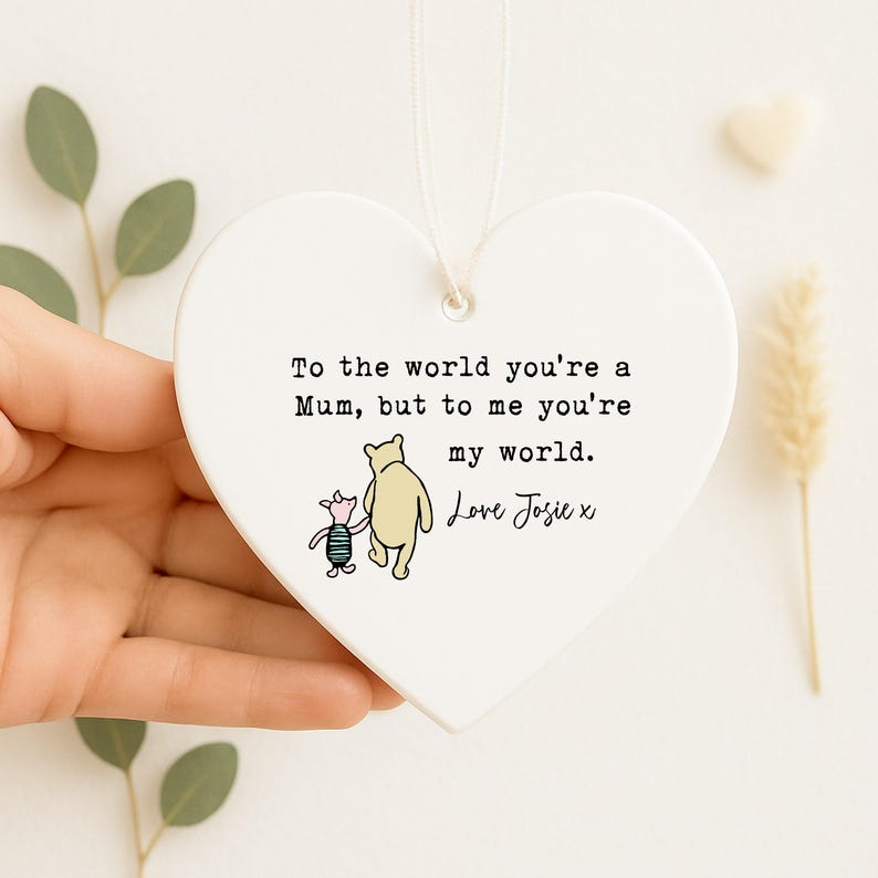 K&ouml;nnte beinhalten: Ein herzf&ouml;rmiger, wei&szlig;er Keramikschmuck mit Aufh&auml;ngeschnur. Er zeigt eine Illustration von Winnie Puuh und Ferkel mit dem Text: "To the world you're a Mum, but to me you're my world. Love Josie x."