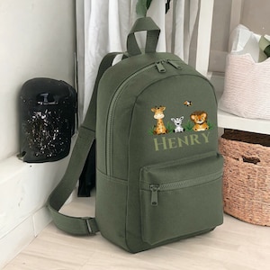 May include: An olive green backpack with a front zip pocket and adjustable straps. It features an embroidered design of a giraffe, zebra, and lion, with the name "HENRY" below. The backpack has a top handle and a double zip closure.