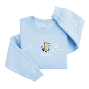 May include: Light blue sweatshirt with a white graphic of Winnie the Pooh and Piglet with the text "Mama Bear Est. 2024". The right sleeve has the text "Jaden Olivia" with a heart.