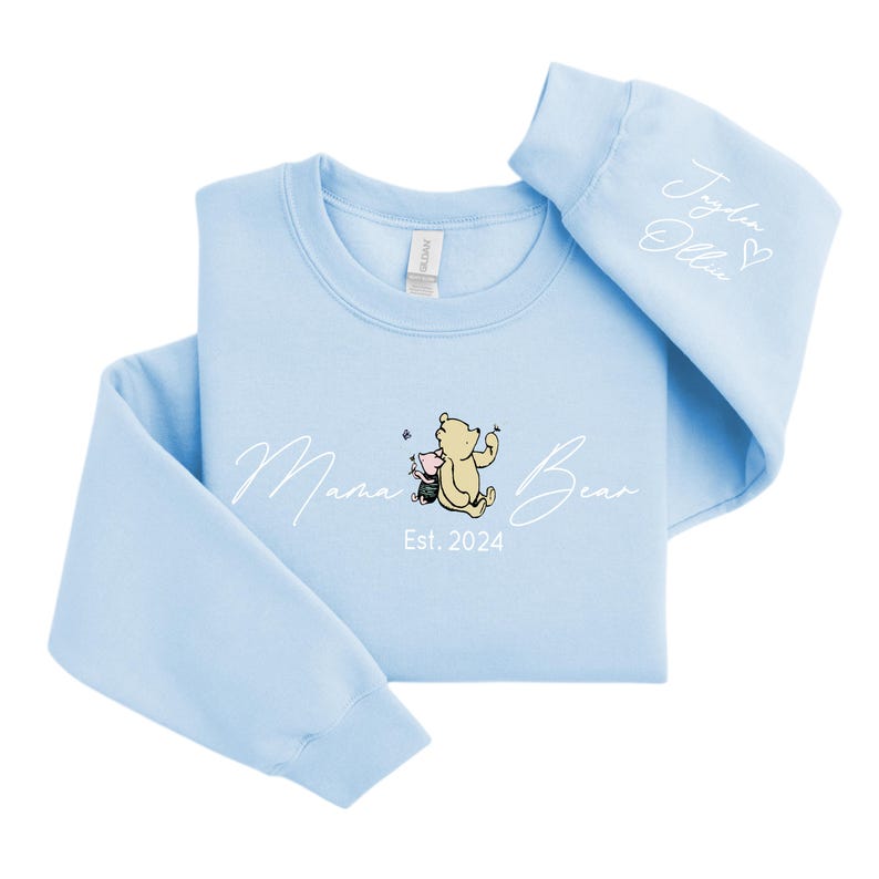 May include: Light blue sweatshirt with a white graphic of Winnie the Pooh and Piglet with the text "Mama Bear Est. 2024". The right sleeve has the text "Jaden Olivia" with a heart.