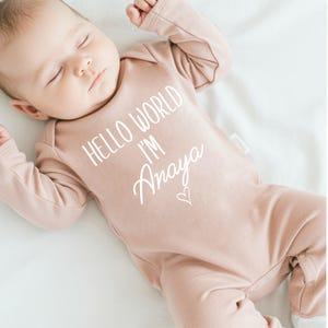 Pode incluir: Um macac&atilde;o rosa para beb&ecirc; com o texto "Hello World I'm Amaya" em branco. Um pequeno cora&ccedil;&atilde;o &eacute; desenhado ao lado do nome.