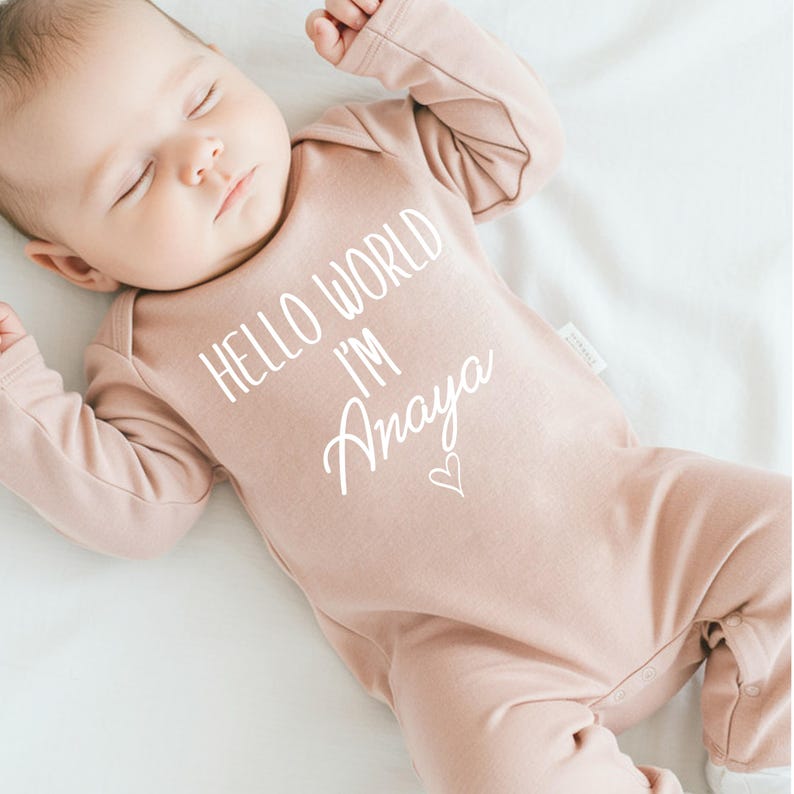Pode incluir: Um macac&atilde;o rosa para beb&ecirc; com o texto "Hello World I'm Amaya" em branco. Um pequeno cora&ccedil;&atilde;o &eacute; desenhado ao lado do nome.