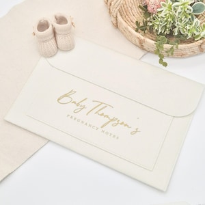 May include: A white leather folder with the text "Baby Thompson's Pregnancy Notes" in gold lettering.