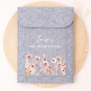 May include: A pale grey felt book folder with a flap closure. The folder has the text "Louise's RED BOOK FOLDER" in white script. A floral design with pink and peach flowers decorates the bottom of the folder.