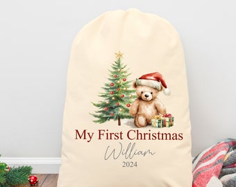 May include: Cream-colored Christmas gift bag featuring a watercolor illustration of a teddy bear wearing a Santa hat next to a decorated Christmas tree. Text reads "My First Christmas" and a name, with the year 2024.