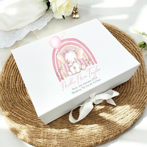 May include: White gift box with a pink and yellow rainbow and a pink elephant with a pink bow. The box is decorated with the text "Millie Mia Taylor" and "Born 3rd January 2024 Weighing 7lb 3oz At 9:23am".