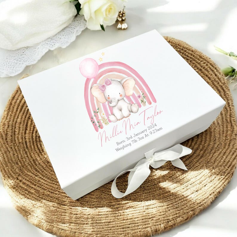 May include: White gift box with a pink and yellow rainbow and a pink elephant with a pink bow. The box is decorated with the text "Millie Mia Taylor" and "Born 3rd January 2024 Weighing 7lb 3oz At 9:23am".