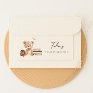 May include: Off-white envelope with a flap, showcasing a watercolour illustration of a teddy bear, books, and a backpack. The text "Talia's Nursery Creations" is in elegant script. The envelope rests on a round, light brown surface.