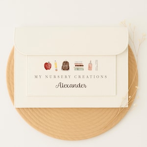 May include: Cream-coloured envelope-style stationery holder with a flap closure. The front features a watercolour illustration of school supplies and the text "MY NURSERY CREATIONS" and "Alexander". The holder is on a round, light brown surface.