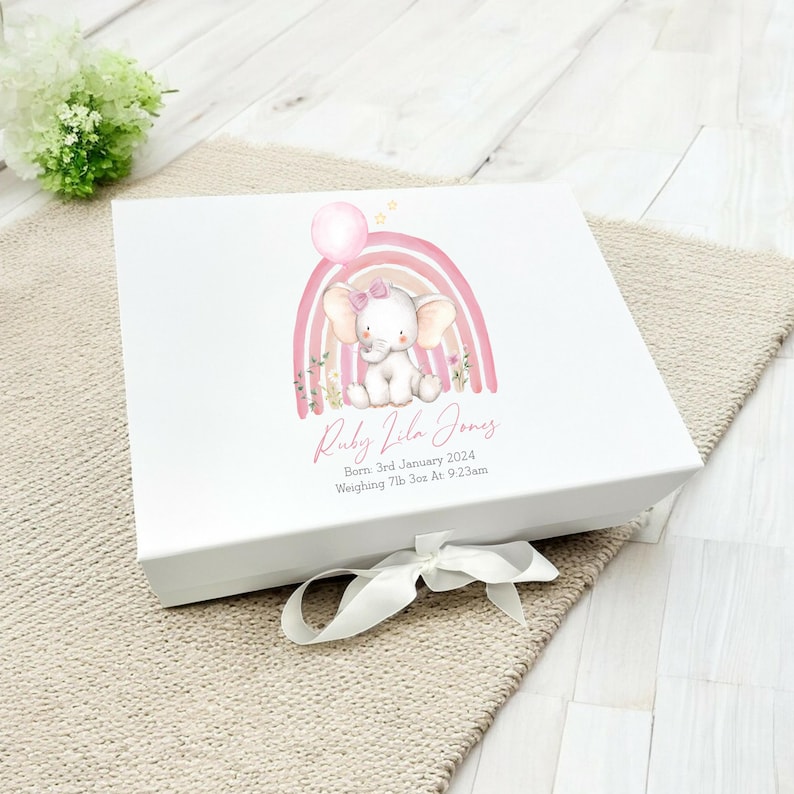 May include: A white keepsake box with a pink and orange rainbow and a pink elephant holding a balloon. The box is decorated with the text "Ruby Vila Jones", "Born: 3rd January 2024", and "Weighing 7lb 3oz At: 9:23am".