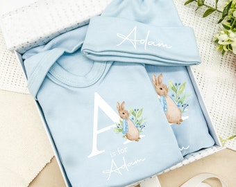 May include: A blue baby onesie with a white letter A and the text "A is for Adam" and a bunny rabbit illustration. A blue hat with the name "Adam" is included in the gift set.