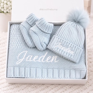 May include: A light blue knitted baby set in a white gift box, including a blanket, two booties, and a beanie with a pom-pom. The blanket and beanie feature the embroidered name Jaeden in white script. The box lid displays allaboutthebump.