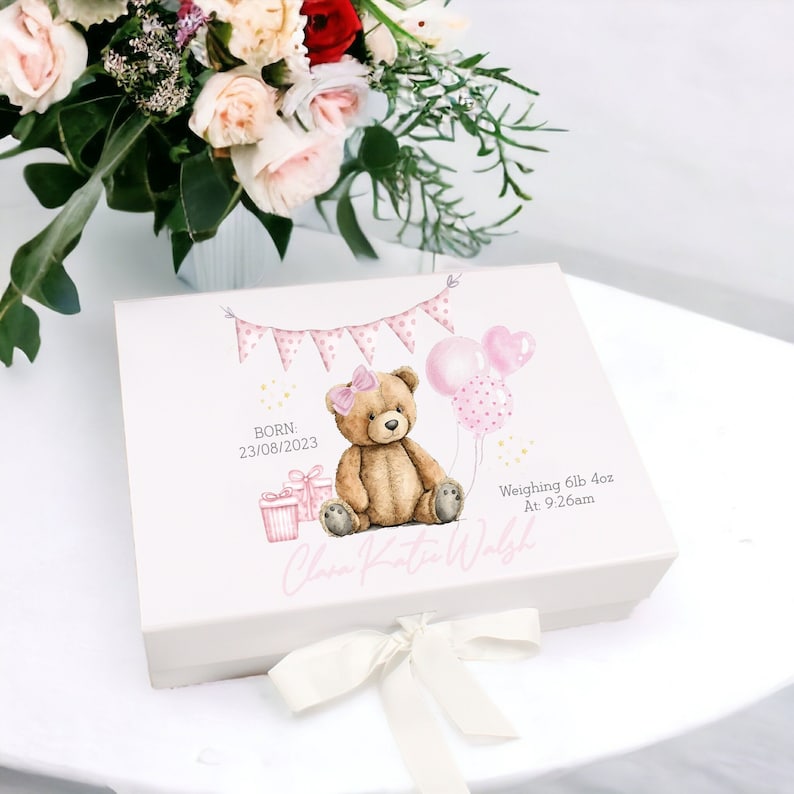 May include: A white keepsake box with a watercolor illustration of a teddy bear, balloons, and gift boxes. The box includes the text "Born: 23/08/2023", "Weighing 6lb 4oz At: 9:26am", and a name. A cream ribbon is tied around the box.