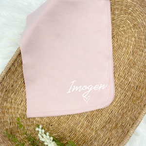 Pode incluir: Um cobertor de beb&ecirc; macio de cor rosa com o nome "Imogen" bordado em branco com um pequeno cora&ccedil;&atilde;o.