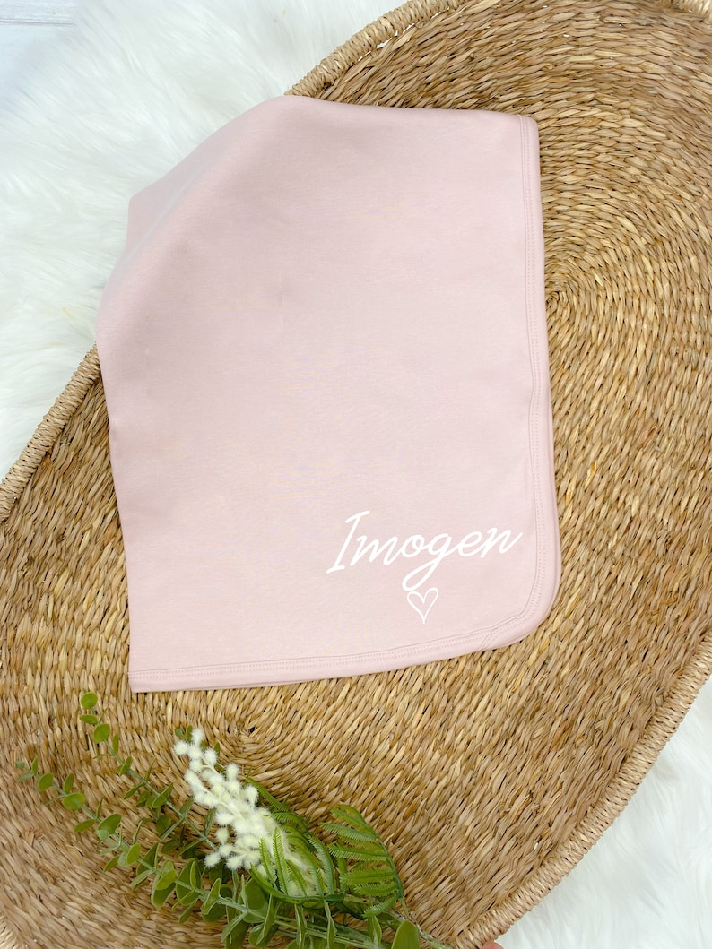 Pode incluir: Um cobertor de beb&ecirc; macio de cor rosa com o nome "Imogen" bordado em branco com um pequeno cora&ccedil;&atilde;o.