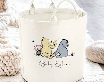 May include: Cream-coloured round storage basket with two handles. The basket features a graphic of Winnie the Pooh, Piglet, and Eeyore. The text "Baby Eglan" is written in a script font below the characters. The basket is on a white rug.