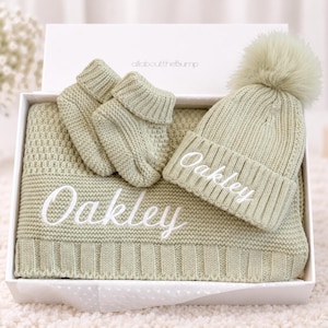 May include: A sage green knitted baby gift set in a white box, including a blanket, a pair of booties, and a pom-pom hat. The blanket and hat are embroidered with the name Oakley in white script. The set features a textured knit pattern.