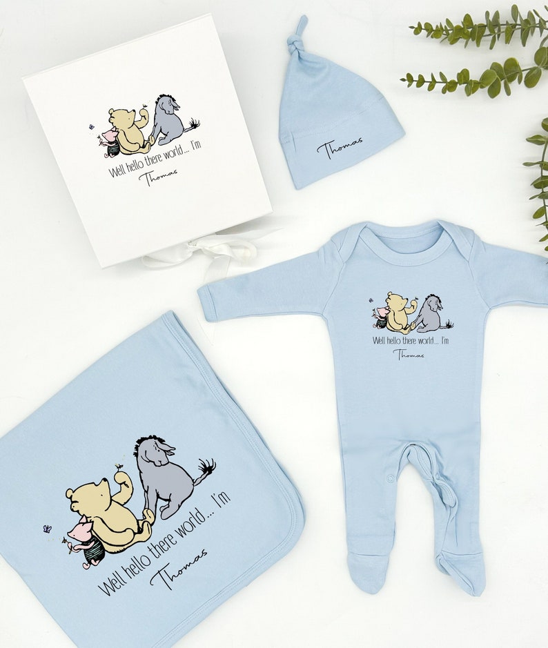 May include: A baby gift set in blue featuring a Winnie the Pooh and Eeyore design. The set includes a onesie, a hat, and a blanket. The text on the items reads "Well hello there world... I'm Thomas".
