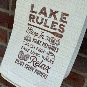 May include: A white kitchen towel with brown embroidery that reads "LAKE RULES" and includes a list of lake rules: "Sleep In", "Make Memories", "Catch Fish", "Take Long Walks", "Relax", and "Enjoy Every Moment".