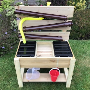 May include: A wooden water table for outdoor play, featuring brown guttering, a yellow flexible tube, and two black trays. Includes a clear plastic jug and a red bucket. Set on a green lawn.