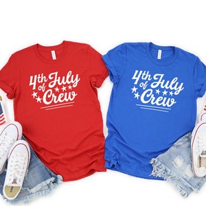 Puede incluir: Dos camisetas, una roja y otra azul, con el texto "4th of July Crew" en blanco. Las camisetas est&aacute;n dispuestas sobre un fondo blanco con unos pantalones cortos de mezclilla azul y zapatillas blancas.