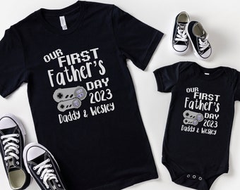 May include: A black t-shirt and a black bodysuit with a white graphic of a video game controller and the text "Our First Father's Day 2023 Daddy & Wesley".