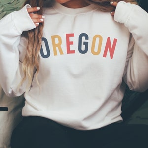 May include: White sweatshirt with the word "OREGON" printed in rainbow colors across the front.