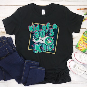 May include: Black t-shirt with the text "Kid of a 90's Kid" in a colorful retro design. The shirt is paired with dark blue jeans and white sneakers. Crayons are scattered around the shirt.
