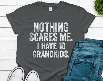 Puede incluir: Una camiseta gris oscuro con el texto "Nothing scares me. I have 10 grandkids." impreso en blanco. La camiseta está colocada sobre una superficie de madera blanca con un par de zapatillas blancas y unos vaqueros azules.