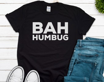 Könnte beinhalten: Schwarzes T-Shirt mit weißem Text, der "BAH HUMBUG" lautet. Das Shirt liegt flach auf einer weißen Holzoberfläche mit einem Paar weißen Sneakers und einer blauen Jeans.