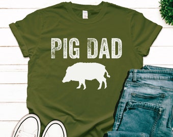 Könnte beinhalten: Ein grünes T-Shirt mit dem Text "PIG DAD" in weißen, abgenutzten Buchstaben. Unter dem Text befindet sich eine weiße Silhouette eines Wildschweins.