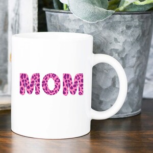 Può includere: Tazza da caffè in ceramica bianca con un design a stampa leopardata rosa e viola che dice "MOM" in grandi lettere.
