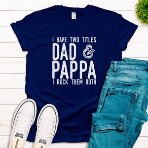 Könnte beinhalten: Ein marineblaues T-Shirt mit weißem Text, der lautet: "Ich habe zwei Titel Papa & Pappa Ich rocke sie beide".