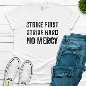 May include: White t-shirt with black text that reads "Strike First Strike Hard No Mercy". The shirt is laid flat on a white wooden surface with a pair of white sneakers and blue jeans.