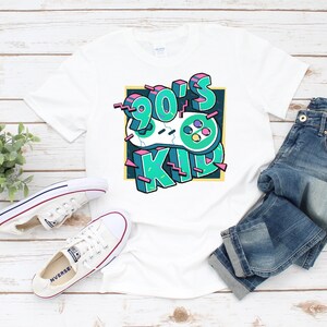 May include: White t-shirt with a graphic design of a retro video game controller and the text "90's Kid" in a colorful, 3D style.