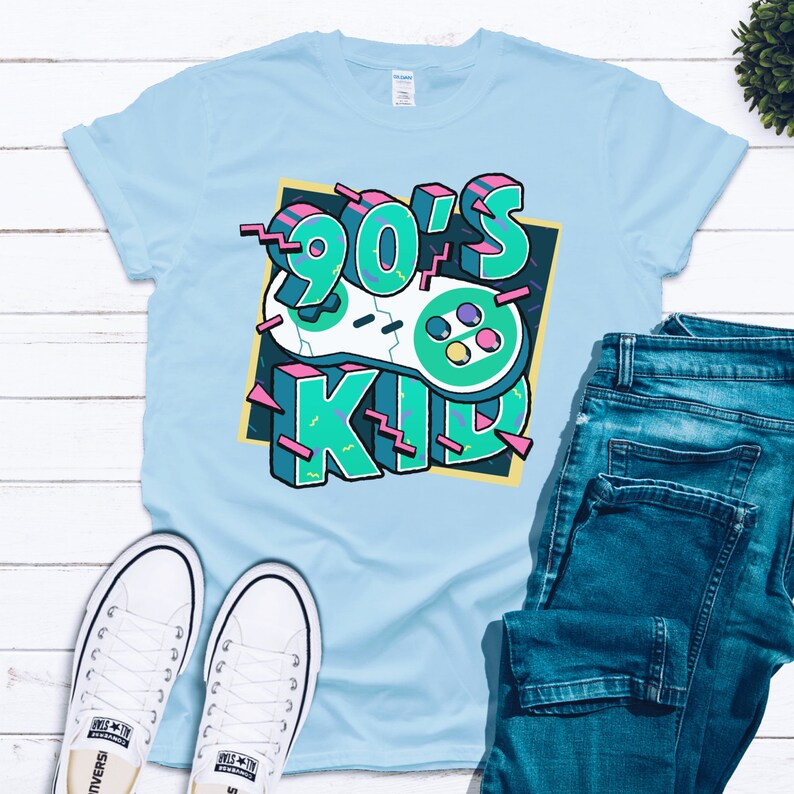 May include: A light blue t-shirt with a graphic design of a retro video game controller and the text "90's Kid" in a colorful, 90s-inspired font.
