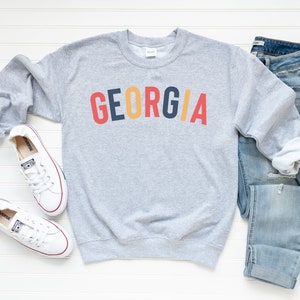 May include: Grey sweatshirt with the word "GEORGIA" printed in red, yellow, blue and black letters. The sweatshirt is laid flat on a white surface with a pair of white trainers and blue jeans.