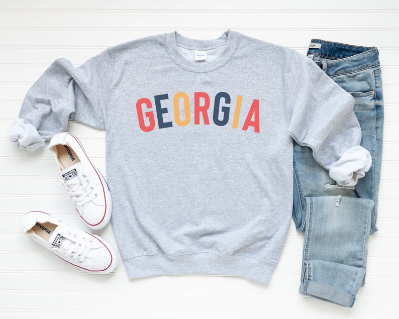 May include: Grey sweatshirt with the word "GEORGIA" printed in red, yellow, blue and black letters. The sweatshirt is laid flat on a white surface with a pair of white trainers and blue jeans.