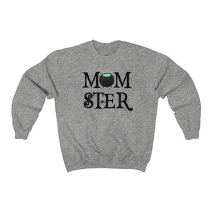 May include: Gray sweatshirt with the text "MOMSTER" in black. The "O" in "MOM" is replaced with a black cauldron with green liquid inside. A small black spider hangs from the cauldron.
