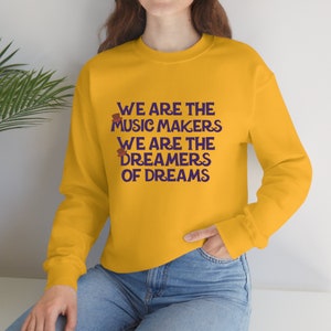 Puede incluir: Una sudadera amarilla con texto morado que dice "We are the music makers. We are the dreamers of dreams". Hay dos pequeños sombreros marrones y morados sobre el texto.